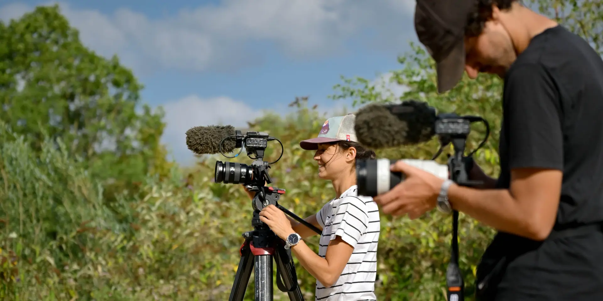Two videographers with cameras film in a tree-lined natural setting under a blue sky.