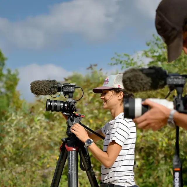 Two videographers with cameras film in a tree-lined natural setting under a blue sky.