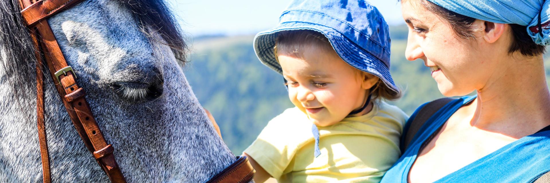 Un jeune enfant porté par sa mère caresse la tête d’un cheval dans une ambiance paisible et ensoleillée à Montcodiol.