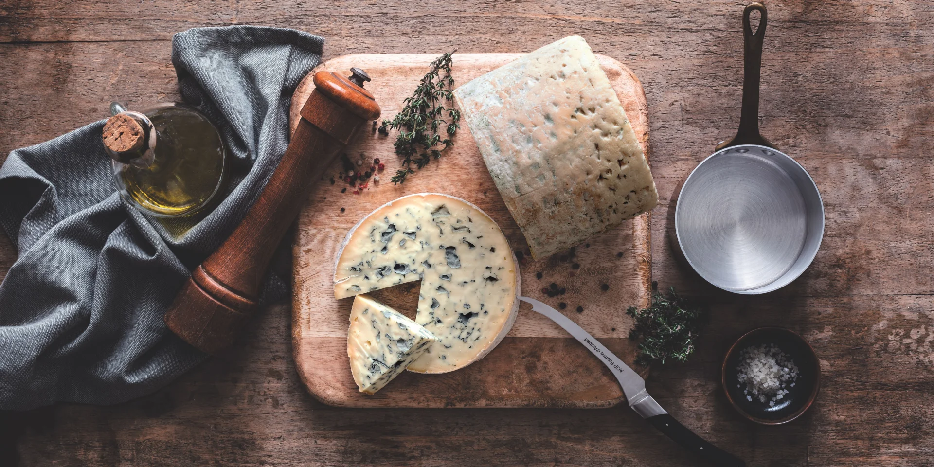 Fromage Fourme d’Ambert tranché sur une planche avec huile d’olive, poivre, thym, sel et ustensiles de cuisine sur table en bois.