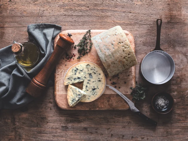 Fromage Fourme d’Ambert tranché sur une planche avec huile d’olive, poivre, thym, sel et ustensiles de cuisine sur table en bois.