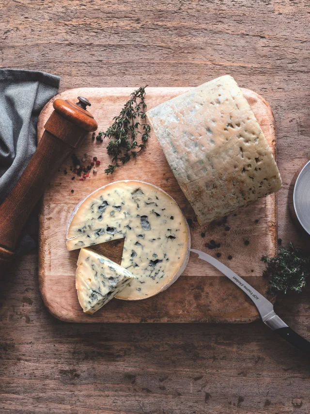 Fromage Fourme d’Ambert tranché sur une planche avec huile d’olive, poivre, thym, sel et ustensiles de cuisine sur table en bois.