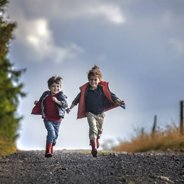 Deux enfants, un garçon et une fille, courent sur une route entourée de sapins et de clôtures en bois.
