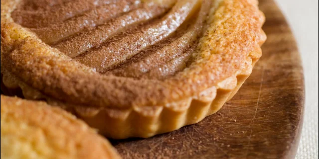 Gros plan sur une tartelette de poire décorative, avec des tranches de poire et une queue de fruit visible, posée sur un support en bois.