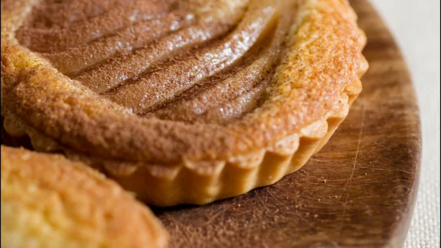 Gros plan sur une tartelette de poire décorative, avec des tranches de poire et une queue de fruit visible, posée sur un support en bois.