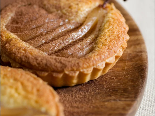Gros plan sur une tartelette de poire décorative, avec des tranches de poire et une queue de fruit visible, posée sur un support en bois.