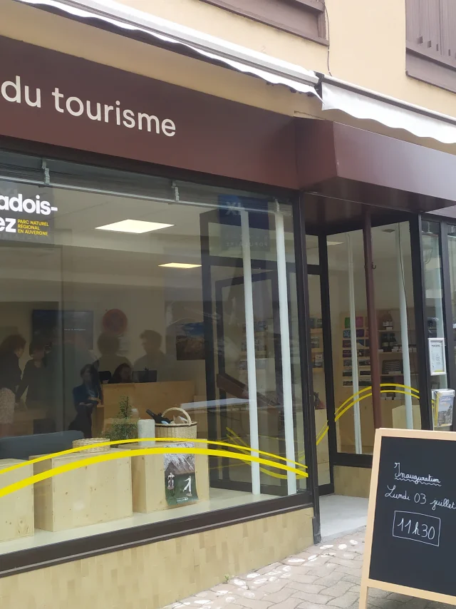 Maison du Tourisme d'Ambert, façade avec la marque et le nom visibles, rue avec des passants et la pharmacie à côté.