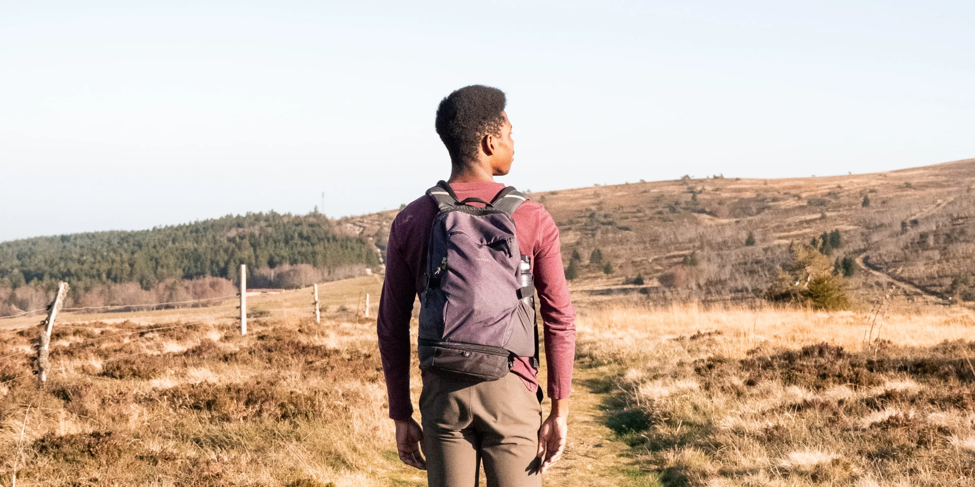 Un homme de dos, équipé d’un sac à dos, marche sur un sentier au cœur d’un paysage naturel vallonné et ensoleillé.