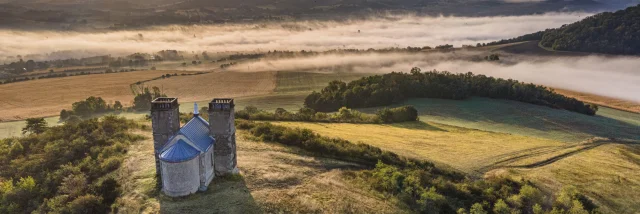 Vue aérienne du site des Turlurons, une chapelle en pierre flanquée de deux tours, située sur une colline. En arrière-plan, un paysage vallonné recouvert de nappes de brume sous la lumière dorée du lever du soleil.