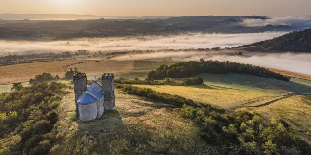 Vue aérienne du site des Turlurons, une chapelle en pierre flanquée de deux tours, située sur une colline. En arrière-plan, un paysage vallonné recouvert de nappes de brume sous la lumière dorée du lever du soleil.