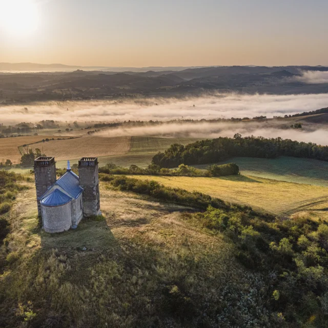 Vue aérienne du site des Turlurons, une chapelle en pierre flanquée de deux tours, située sur une colline. En arrière-plan, un paysage vallonné recouvert de nappes de brume sous la lumière dorée du lever du soleil.