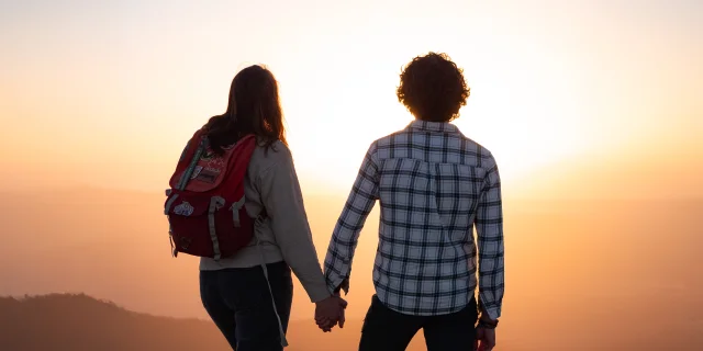 Couple de randonneurs vu de dos, se tenant la main face à un coucher de soleil à Peyre Mayou dans le Livradois-Forez sur les montagnes, dans un décor naturel au sommet d’une colline ou d’une montagne.