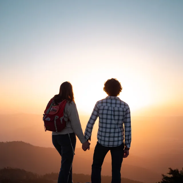 Couple de randonneurs vu de dos, se tenant la main face à un coucher de soleil à Peyre Mayou dans le Livradois-Forez sur les montagnes, dans un décor naturel au sommet d’une colline ou d’une montagne.