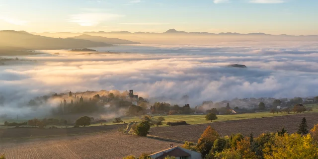 Paysage du Livradois-Forez au lever du jour, avec un village baigné de brume et les volcans d’Auvergne en arrière-plan sous un ciel doré