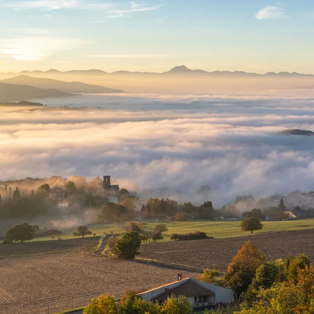 Paysage du Livradois-Forez au lever du jour, avec un village baigné de brume et les volcans d’Auvergne en arrière-plan sous un ciel doré