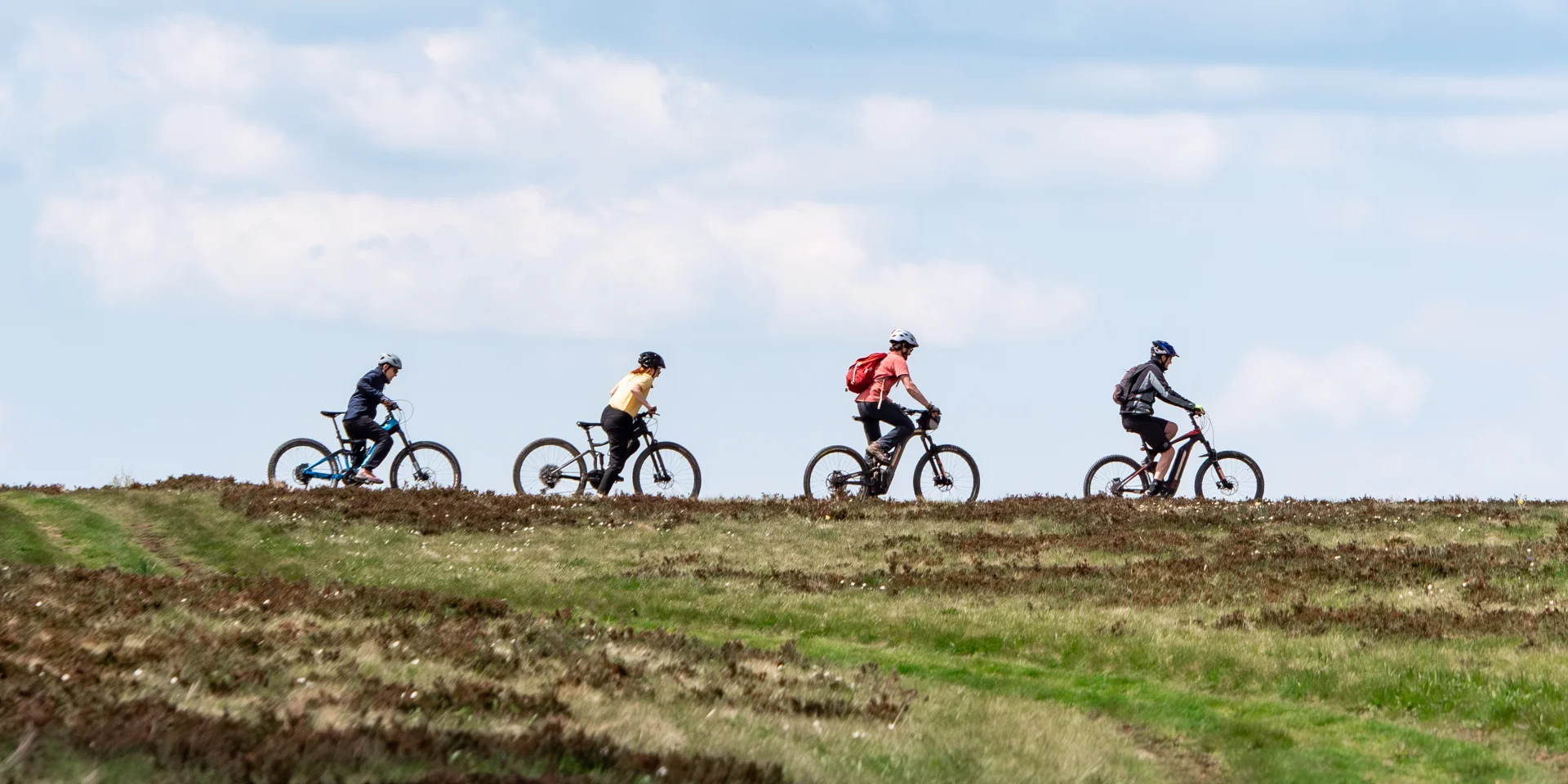 Quatre personnes à vélo s’arrêtent dans un champ en altitude, admirant un paysage de montagnes verdoyantes sous un ciel bleu parsemé de nuages.