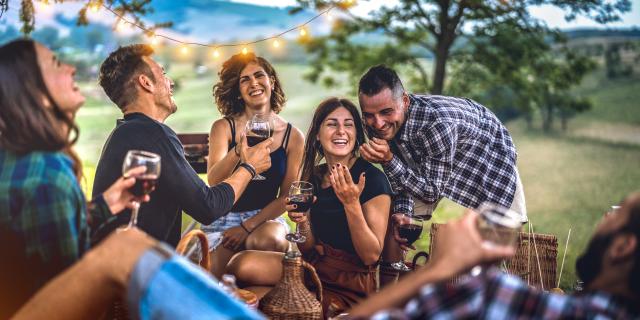 Young friends having fun at vineyard after sunset - Happy people millennial camping at open air pic nic under bulb lights - Youth friendship concept with guys and girls drinking wine at barbeque party