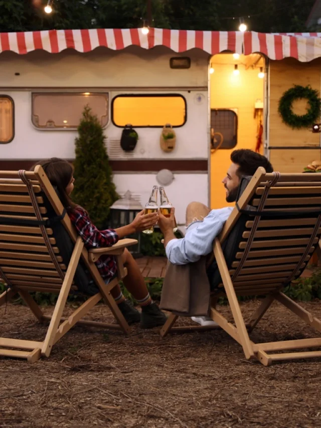 Young couple toasting with bottles of beer near trailer. Camping season