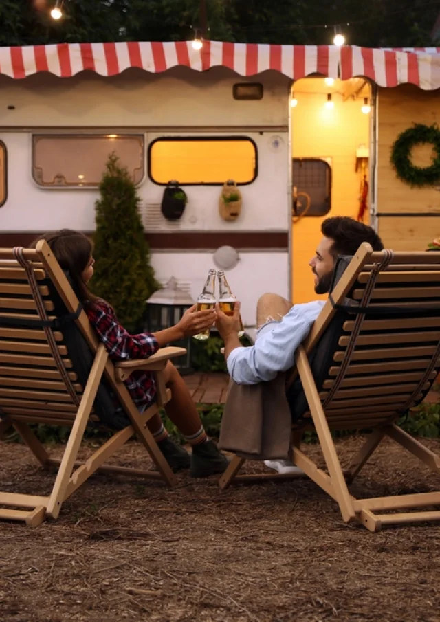 Pareja joven brindando con botellas de cerveza cerca del remolque. Temporada de camping