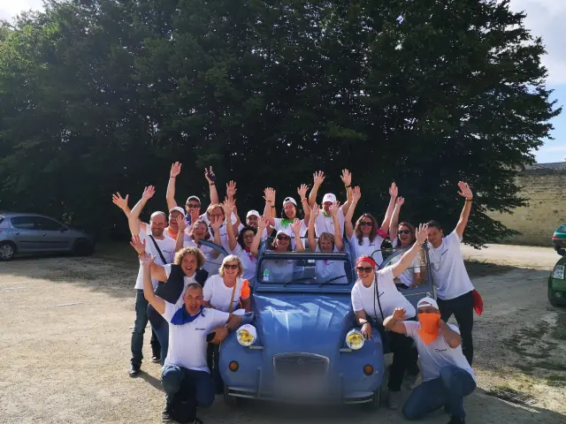 Une grande équipe de professionnels pose joyeusement autour d'une Citroën 2CV bleue