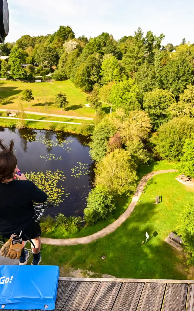 Vue en hauteur, d'une plateforme de départ de tyrolienne, d'un étang paisible entouré de pelouses vertes et d'arbres, prise depuis une plateforme en bois