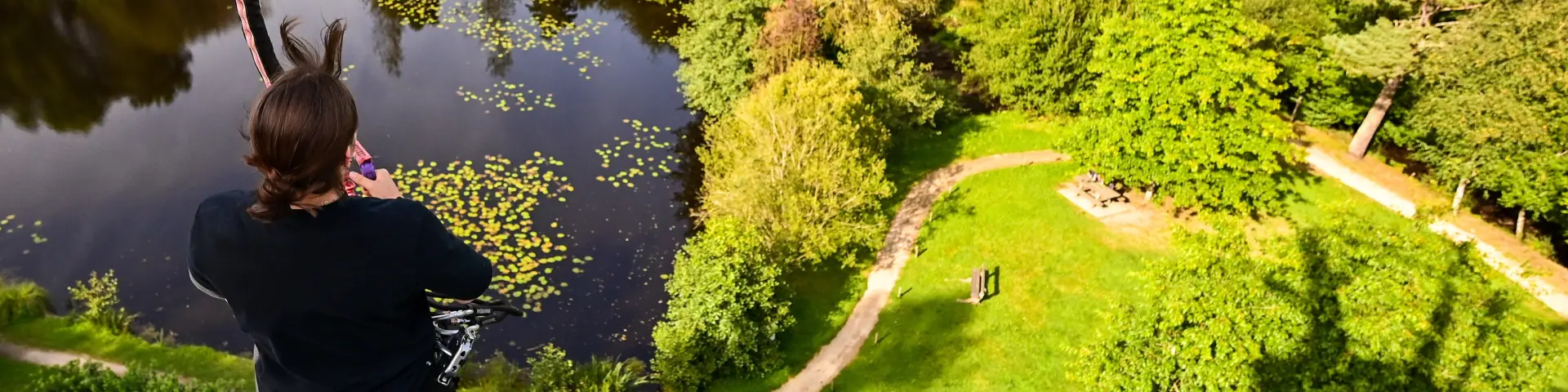 Vue en hauteur, d'une plateforme de départ de tyrolienne, d'un étang paisible entouré de pelouses vertes et d'arbres, prise depuis une plateforme en bois