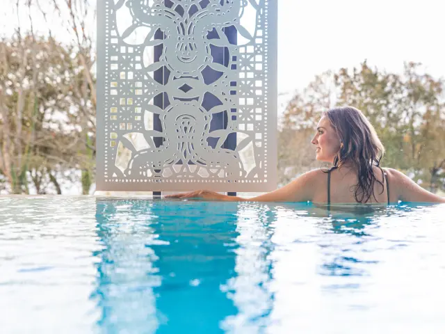 Une femme de profil se détend au bord d'une piscine à débordement, à côté d'une paroi métallique blanche finement sculptée de motifs géométriques