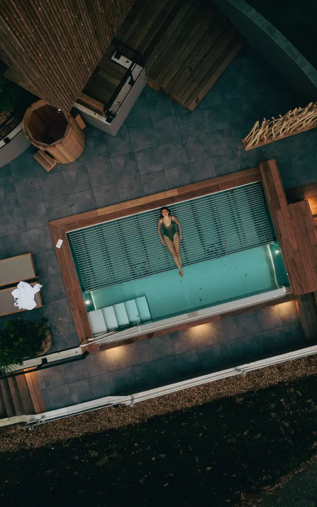 Vue aérienne en plongée totale d'une piscine extérieure illuminée de spa, entourée de lits de repos et de terrasses en bois dans la pénombre
