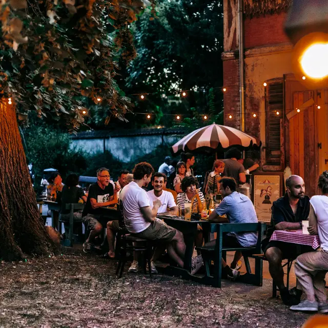 Plusieurs tables en extérieur avec des personnes qui boivent un verre sous des arbres et près d'un vieux bâtiment.