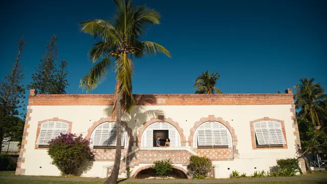 Façade d’un bâtiment historique du bagne de Nouville (CREIPAC) à Nouméa, avec palmiers et volets blancs.