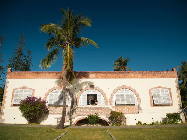 Facade of a historic convict-era building at Nouville (CREIPAC) in Nouméa, with palm trees and white shutters.