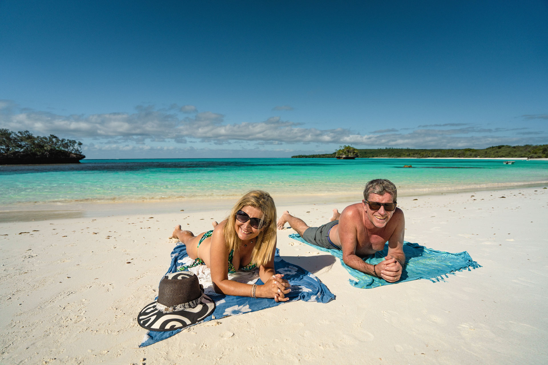 Couple relaxing on towels on a white-sand beach by a turquoise lagoon under a clear blue sky.