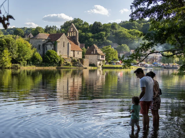 Une famille trempe les pieds dans la rivière Dordogne, avec en arrière-plan le village pittoresque de Beaulieu-sur-Dordogne