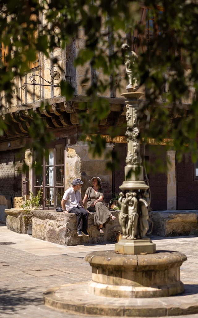 Sur la charmante place du Mercadial à Saint-Céré, un couple est assis, savourant l’ambiance paisible du lieu.