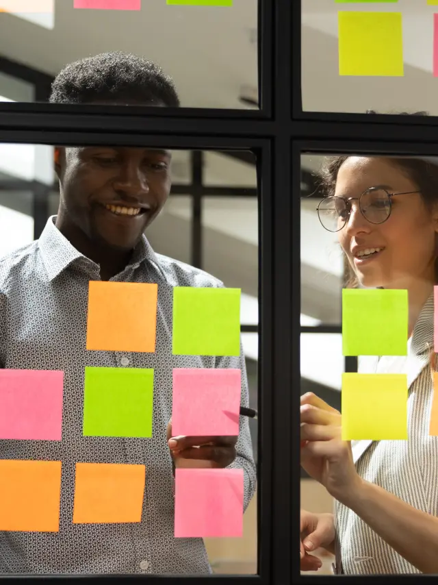 Multiracial colleagues developing business ideas use post-it notes view through glass, mates read written tasks shown on stickers attached on wall, effective way do work don't forget anything concept