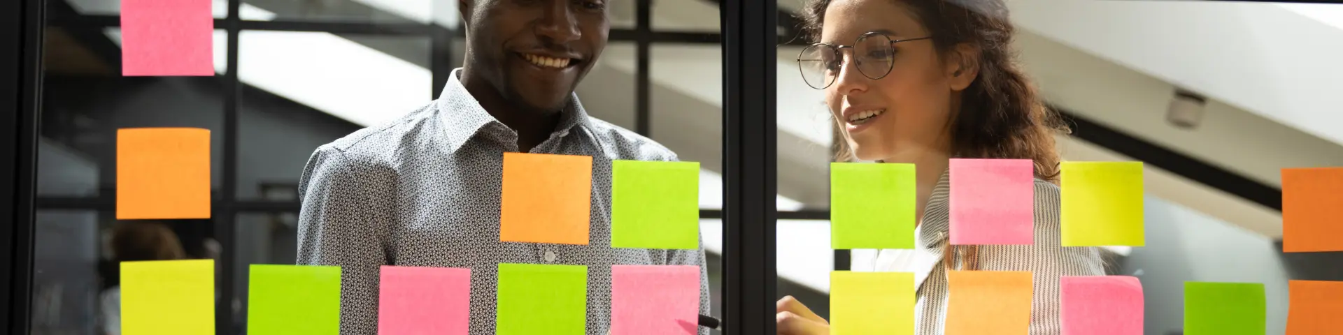 Multiracial colleagues developing business ideas use post-it notes view through glass, mates read written tasks shown on stickers attached on wall, effective way do work don't forget anything concept