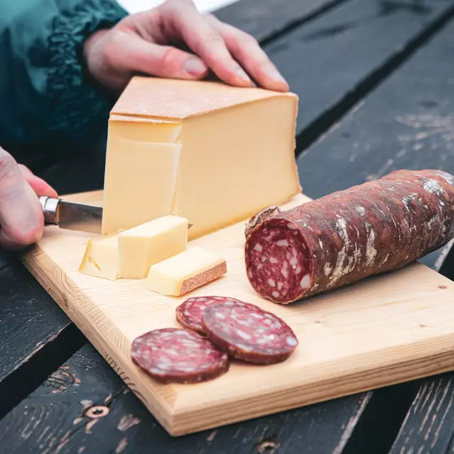 A wooden board supports a pre-cut sausage, while someone cuts slices of Abondance with an Opinel knife.