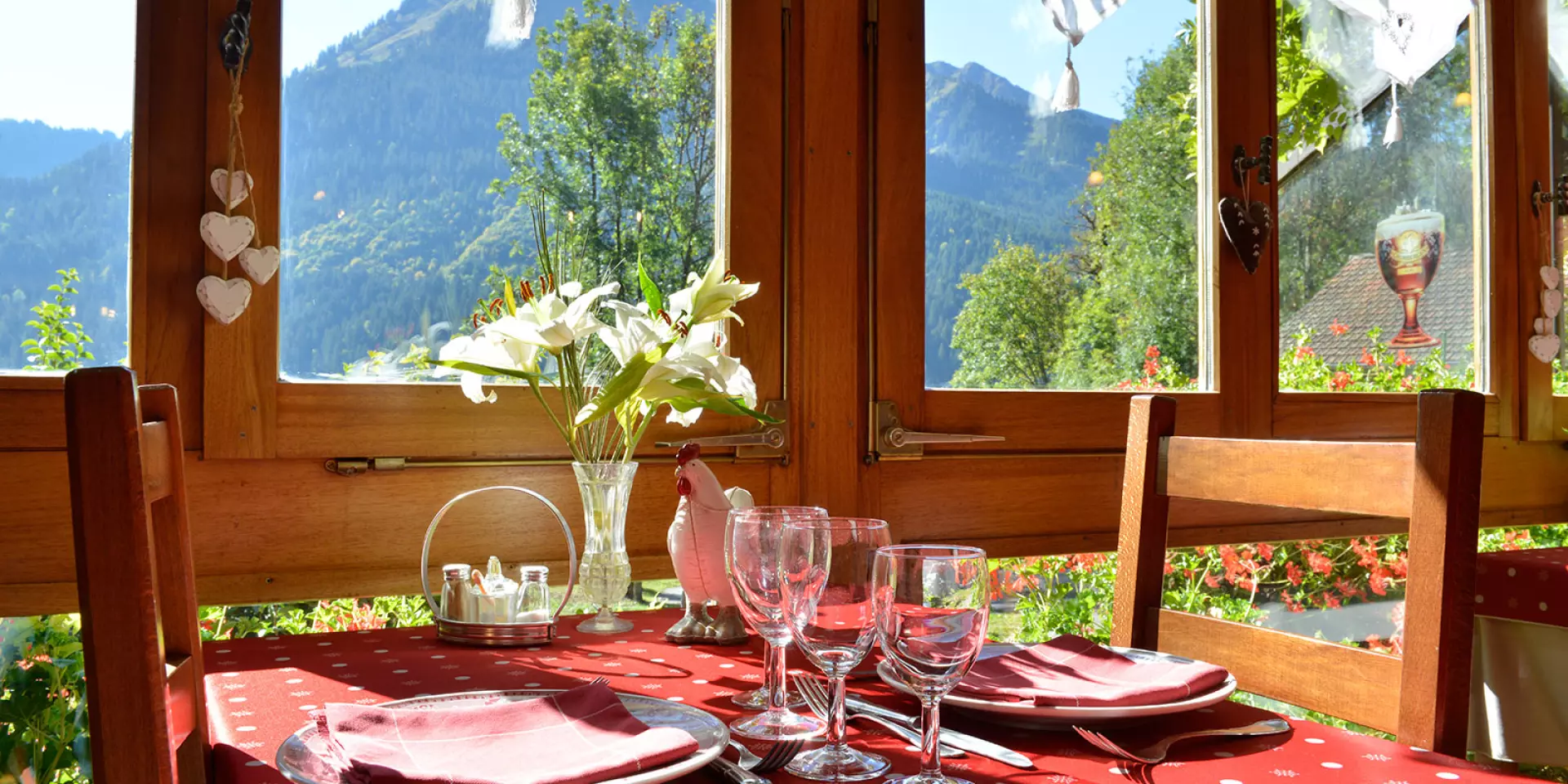 Table prête à servir un repas, nappe rouge, fleurs dans un vase et vue sur la montagne