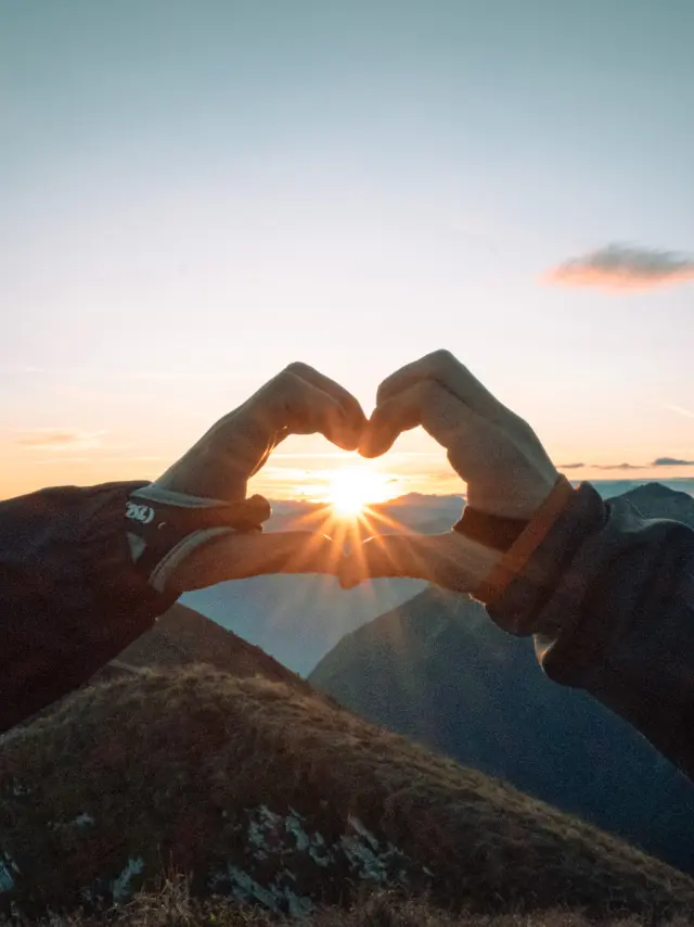 Un coeur fait avec les doigts, entourant le soleil couchant, face aux montagnes.