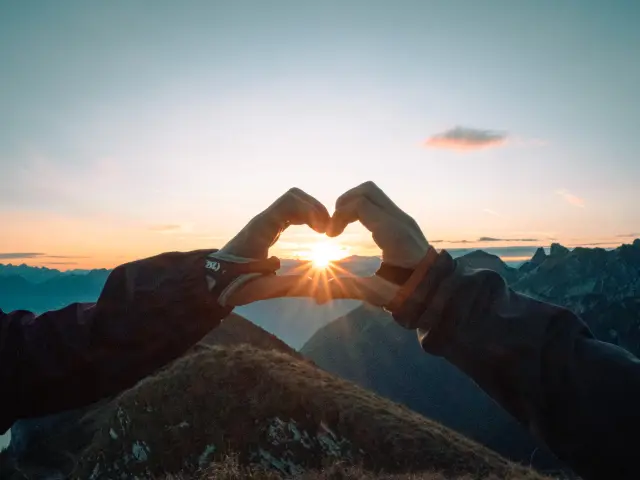 Un coeur fait avec les doigts, entourant le soleil couchant, face aux montagnes.
