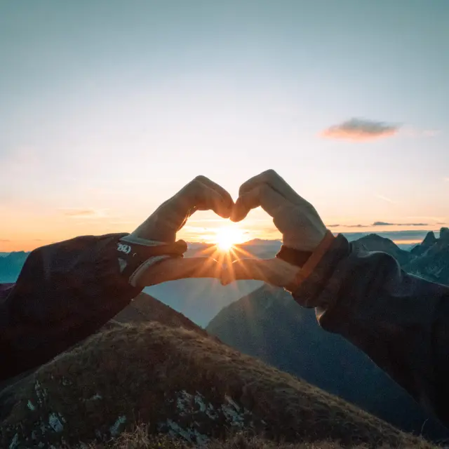 Un coeur fait avec les doigts, entourant le soleil couchant, face aux montagnes.