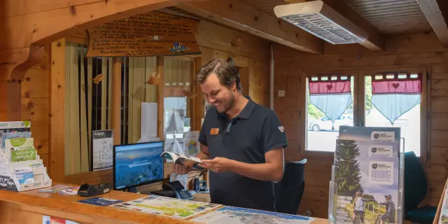 Conseiller de l’Office de Tourisme de Bernex, souriant, derrière le comptoir en bois, présentant une brochure touristique.