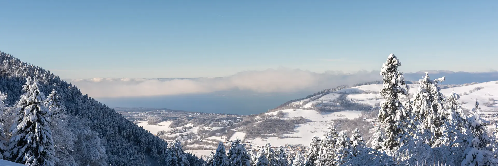 Sur les hauteurs de la station de ski de Bernex, on voit un paysage enneigé avec, en contrebas, une vue sur le Léman et la suisse, de l'autre côté, sous un grand ciel bleu.