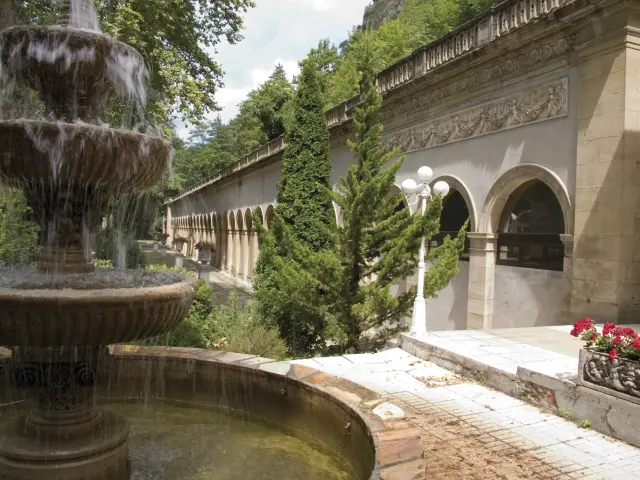 Fuente con varios niveles de agua en un jardín rodeado de vegetación y un edificio antiguo