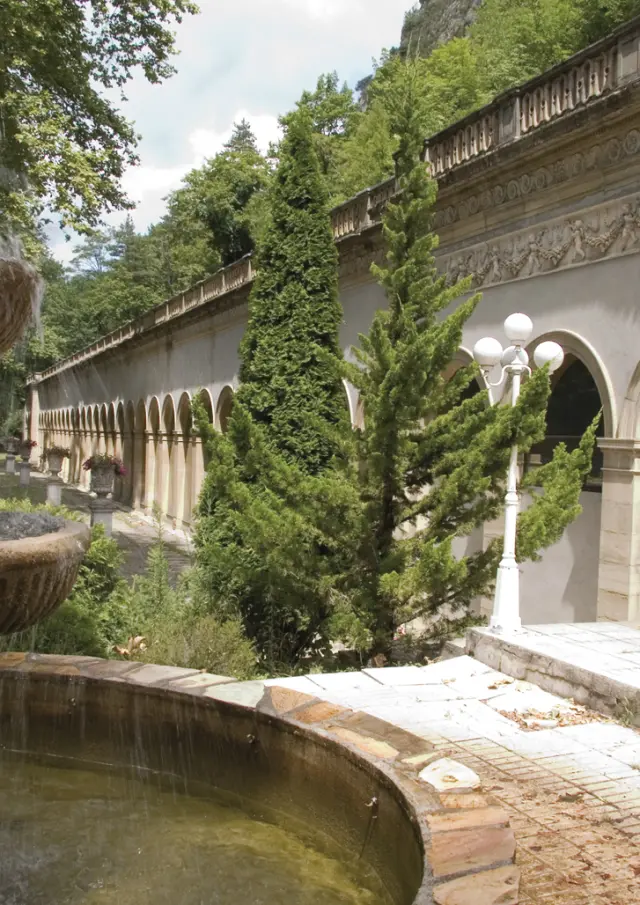 Fountain with multiple water levels in a garden surrounded by vegetation and an old building