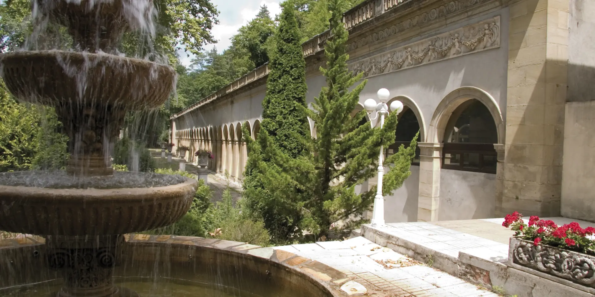Fountain with multiple water levels in a garden surrounded by vegetation and an old building