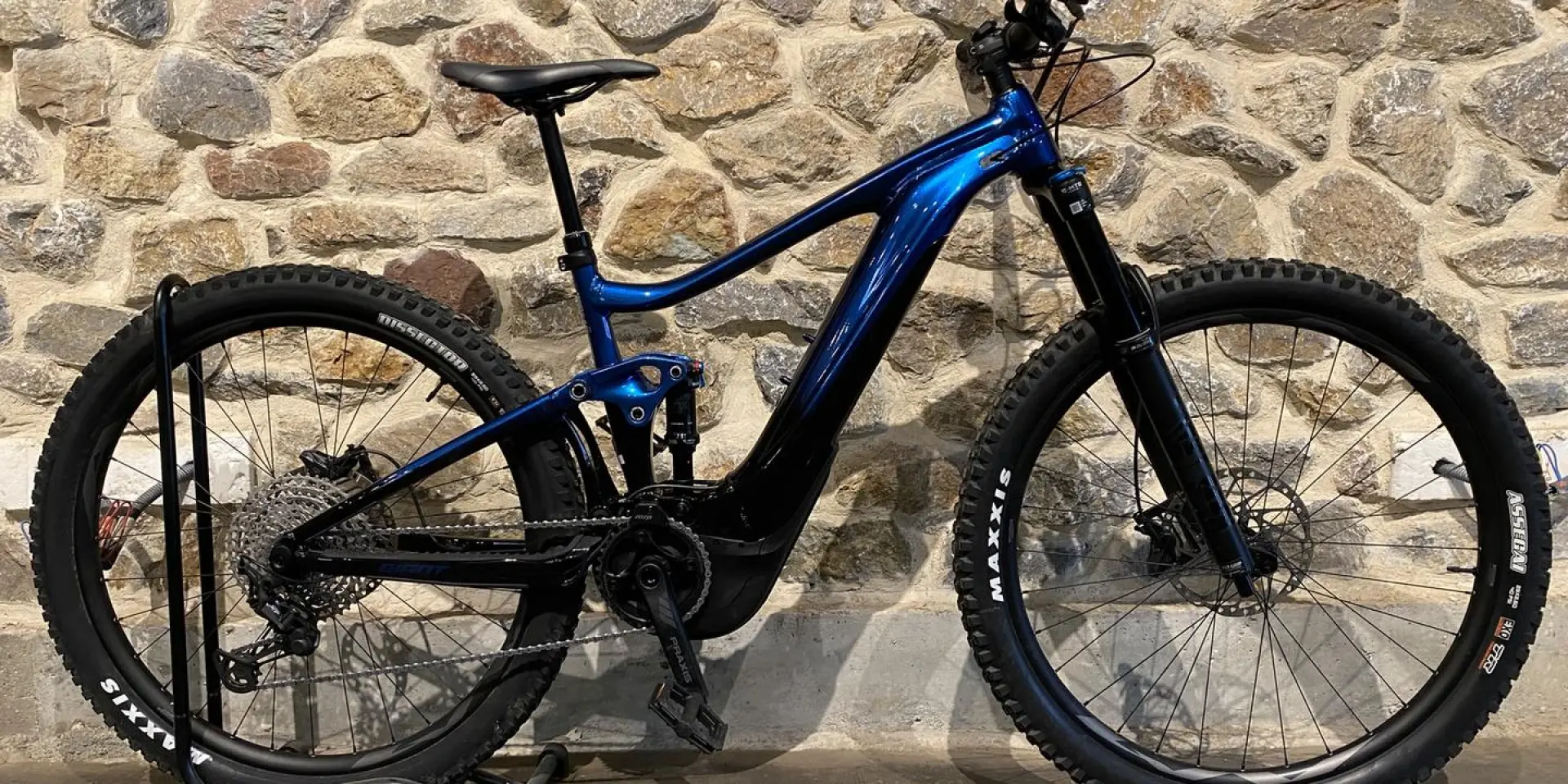 VTT électrique bleu avec des pneus noirs et une suspension avant et arrière, posé contre un mur de pierre