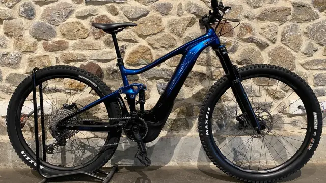 Blue electric mountain bike with black tires and front and rear suspension, leaning against a stone wall
