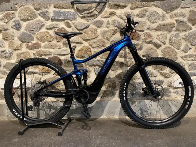 Bicicleta de montaña eléctrica azul con neumáticos negros y suspensión delantera y trasera, apoyada contra una pared de piedra