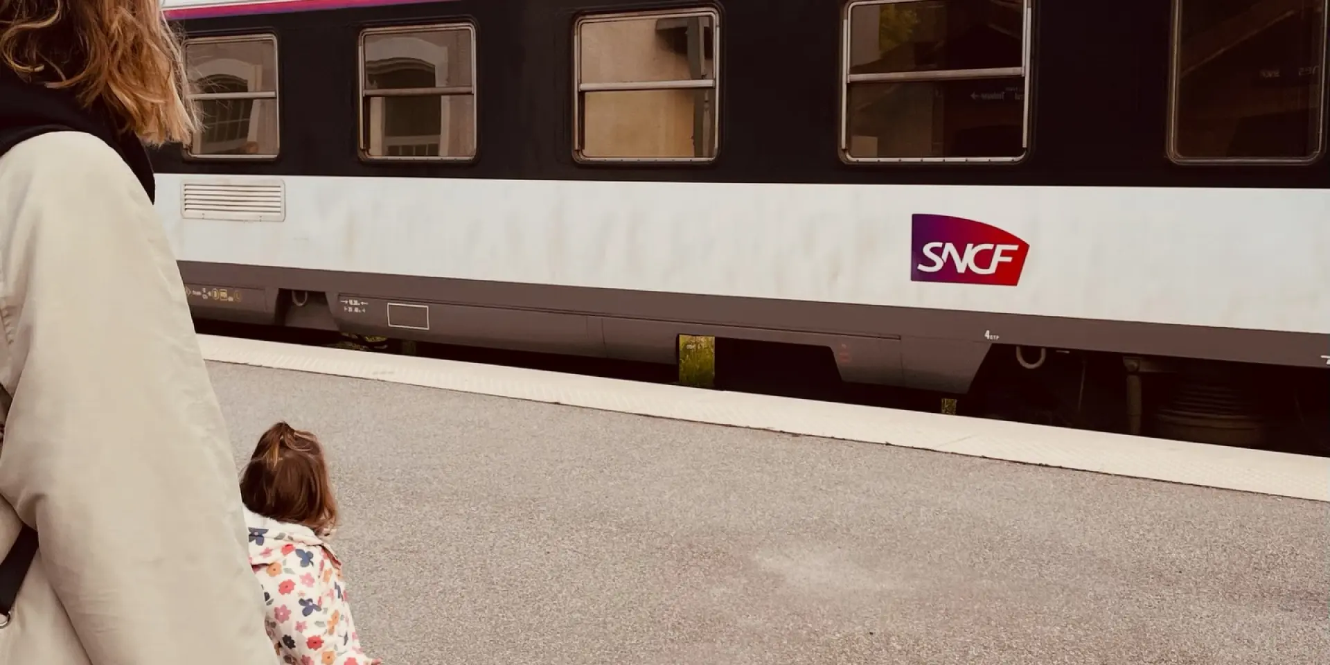 Une femme et un enfant attendent un train de nuit devant un train SNCF avec des montagnes en arrière-plan